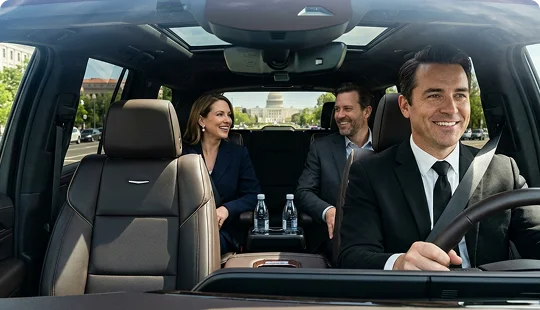 Interior view of a luxury vehicle showing a smiling chauffeur in a suit driving with two passengers chatting in the rear. The passengers enjoy a comfortable ride with water bottles available in the center console as the car travels through the city.