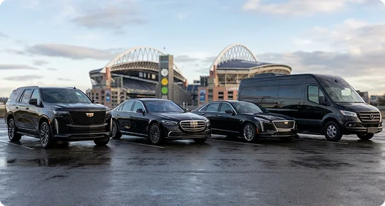 Four black luxury vehicles, including two SUVs, a sedan, and a large sprinter van, are parked in a row in a paved lot with the arched structures of a stadium visible in the background under a cloudy sky.