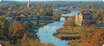 A scenic aerial view of Colonial Williamsburg, Virginia, showing a tranquil river meandering through a historic town with red-brick houses, a stone bridge, and vibrant fall foliage.