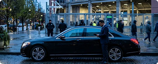 A chauffeur in a dark suit stands beside a black luxury sedan on a cobblestone street, looking down at a tablet, with a stadium entrance and pedestrians in the background.