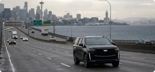 A black Cadillac SUV drives along a multi-lane highway bridge toward the Seattle skyline, with the Space Needle and Elliott Bay visible in the background under an overcast sky.
