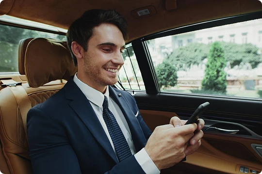A young businessman in a navy blue suit smiles while using his smartphone in the back seat of a premium car with tan leather interior and a cityscape visible outside the window.