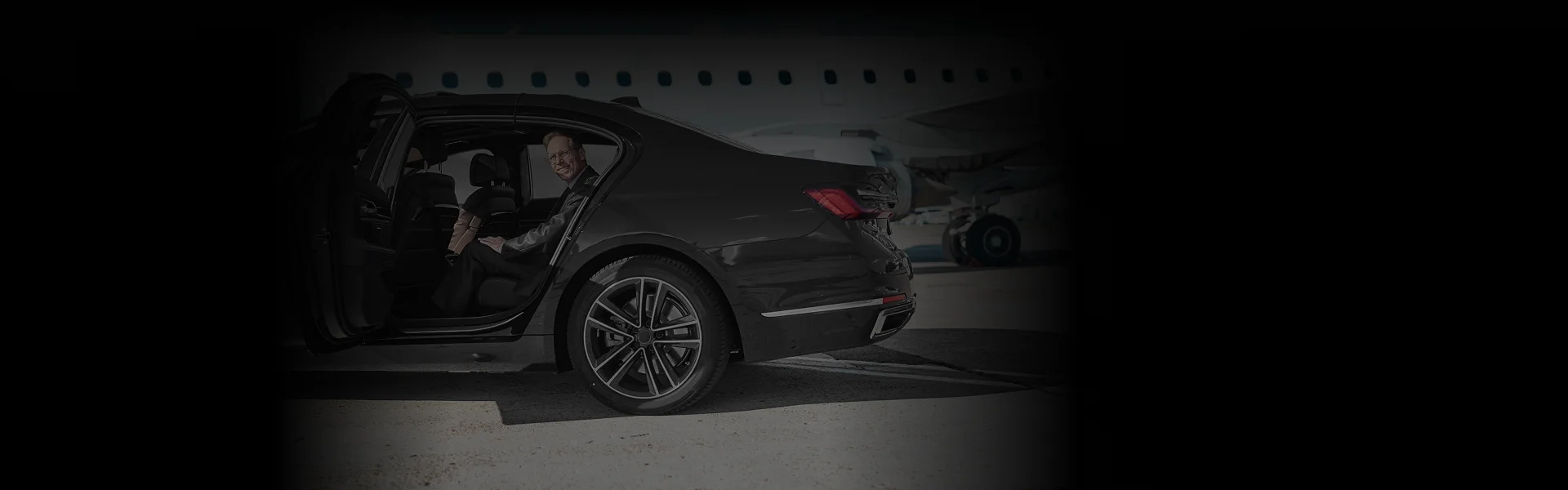 Side-angle view of a luxury black sedan parked near a jet, with a smiling man seated comfortably inside. Captures the premium experience of DCA Airport Car Service with style.